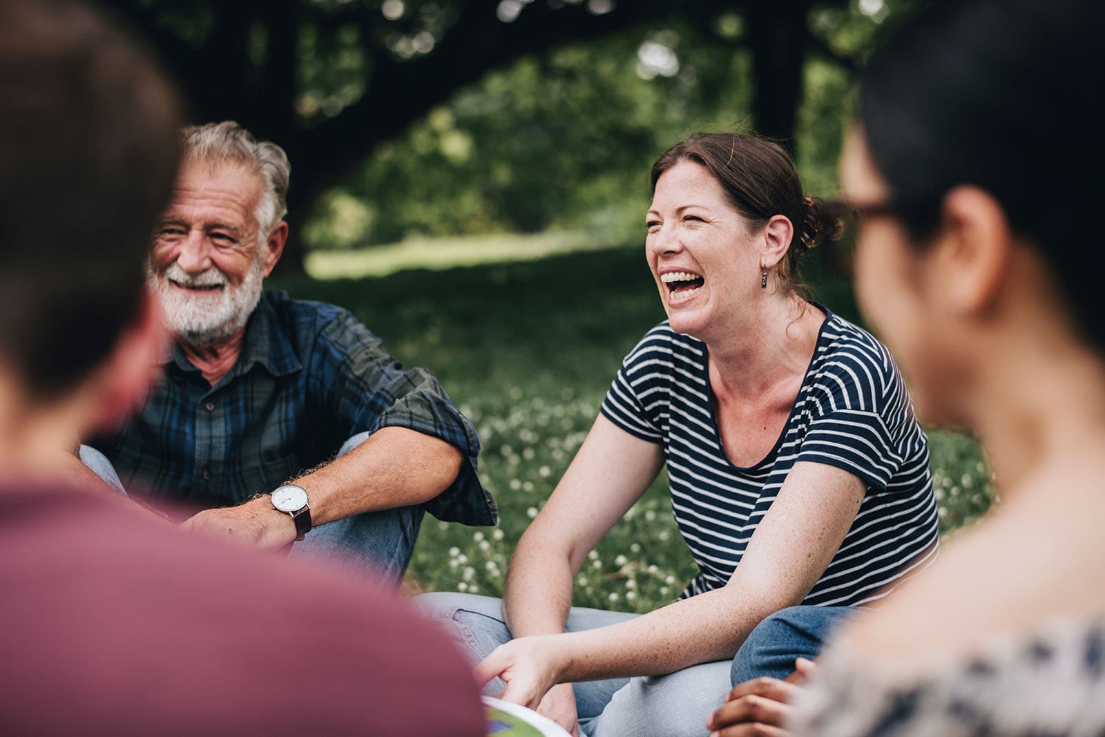 A small group of adults sitting together on the grass in a park, talking and laughing.