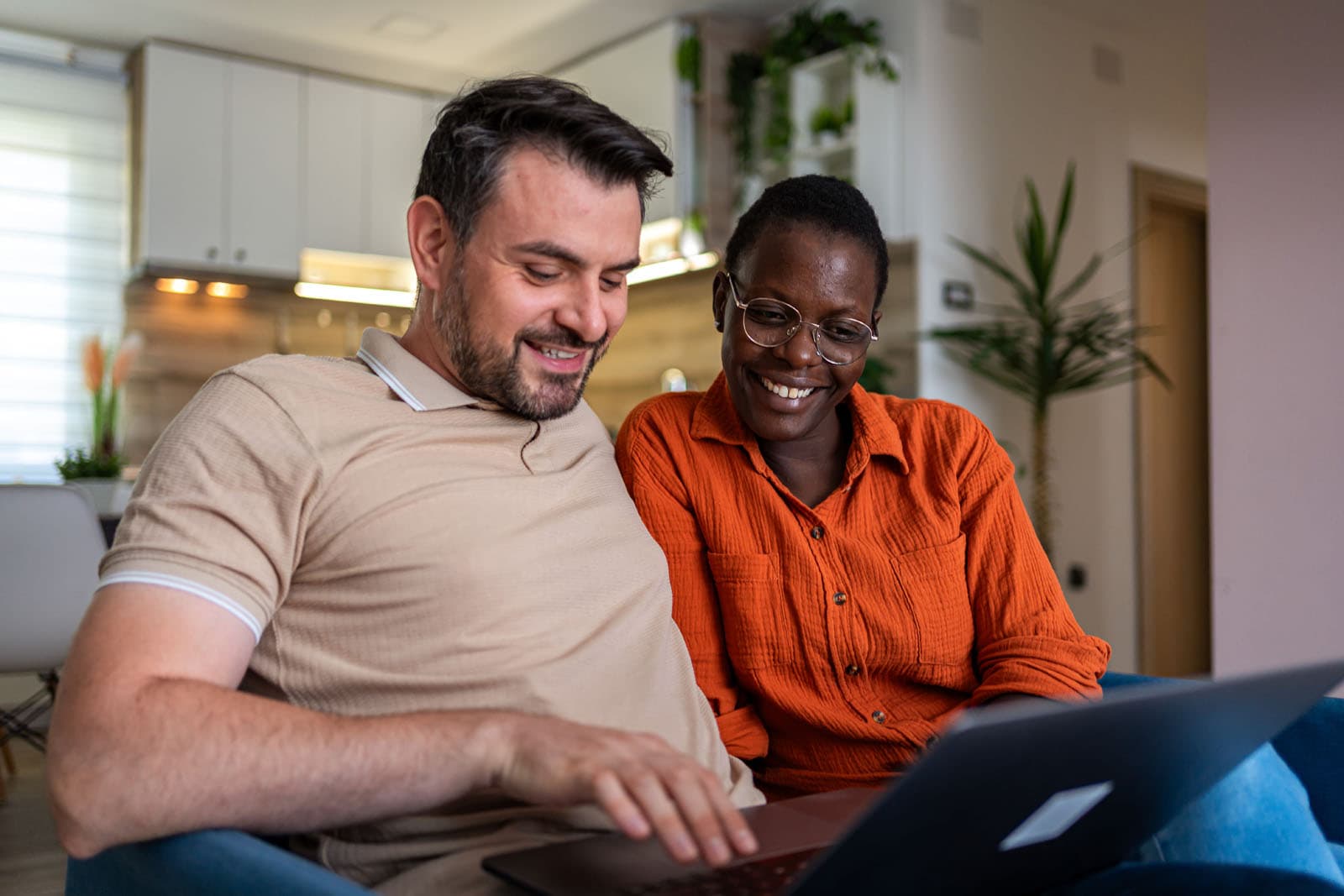 Two adults sitting together at home, smiling as they watch something on a laptop.